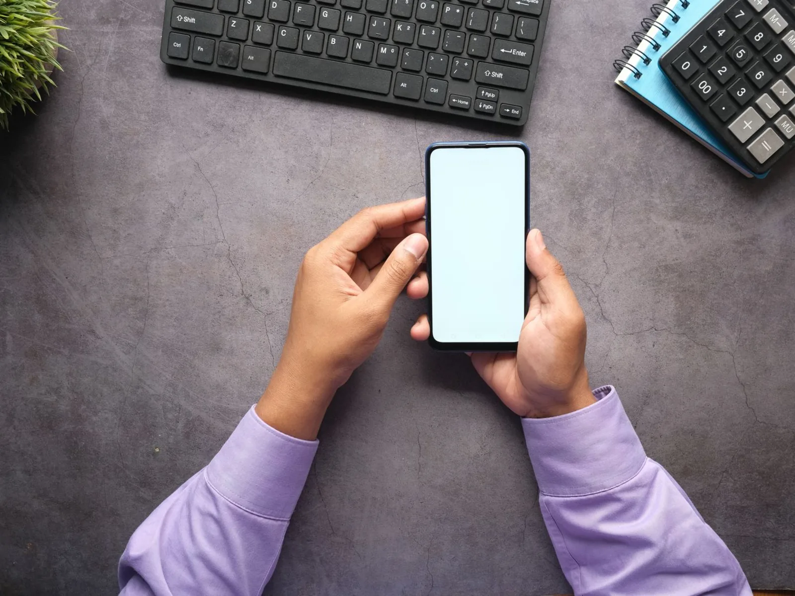 Overhead view of a person holding a smartphone at a desk beside a keyboard and calculator